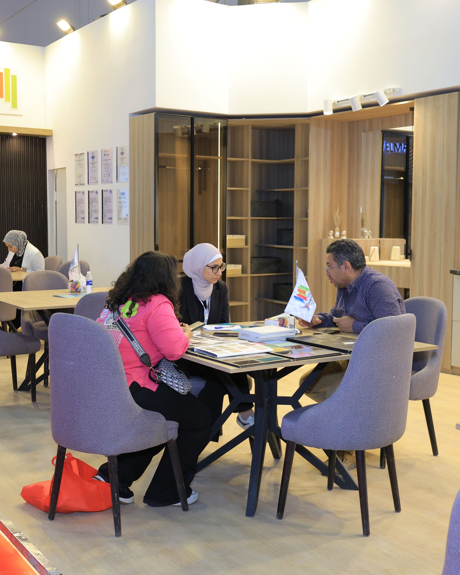 Visitors on the HOMEX floor, 6th edition — seated with an exhibitor inside a full-scale room set, comparing finish samples at the table.